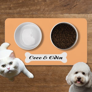 May include: A peach-colored silicone pet feeding mat with two white bowls. One bowl is filled with dry dog food and the other is empty. The mat has a white bone-shaped label with the text "Coco & Odin".