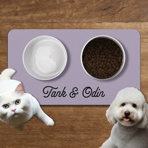 Op de afbeelding: Een paarse siliconen voedermat voor huisdieren met twee witte kommen. Een kom is gevuld met droog hondenvoer en de andere is leeg. De mat heeft de tekst "Tank & Odin" erop gedrukt. Een witte kat en een witte hond kijken naar de kommen.