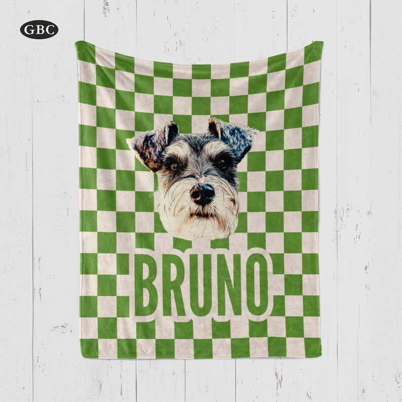 Puede incluir: Una manta a cuadros verdes y blancos con un retrato de perro Schnauzer y el nombre "BRUNO" en letras verdes grandes. La manta tiene un aspecto suave y se muestra sobre un fondo de madera blanca.