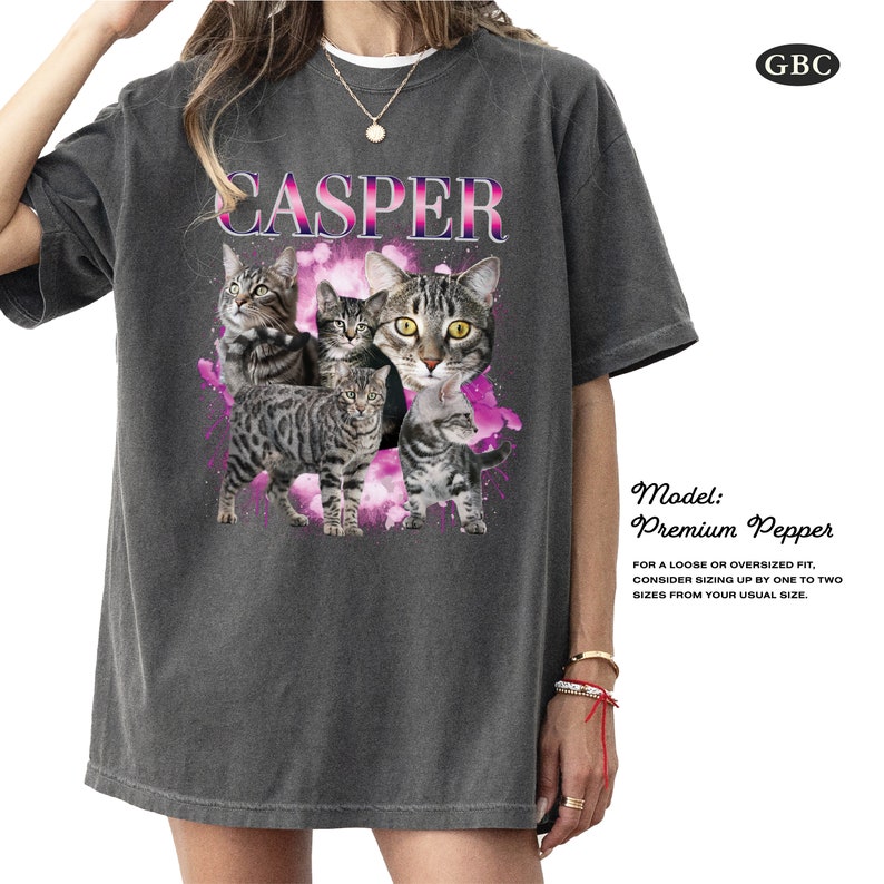 May include: A gray oversized t-shirt with a graphic of four cats in a pink and purple cloud design. The text "CASPER" is printed in white above the cats. The model is wearing the shirt and has a red string bracelet on her wrist.