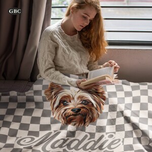 Puede incluir: Una manta a cuadros grises y blancos con una ilustraci&oacute;n detallada de la cara de un Yorkshire Terrier. El nombre del perro, "Maddie", est&aacute; impreso en cursiva en la parte inferior. La manta est&aacute; sobre una persona.