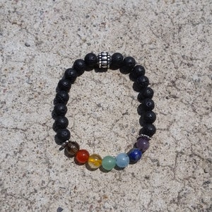 May include: Black lava rock bead bracelet with a silver-colored spacer bead. The bracelet features a row of colorful chakra beads in red, orange, yellow, green, blue, indigo, and violet. The bracelet is on a textured gray surface.