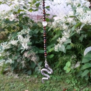 May include: A silver snake charm hangs from a beaded necklace with red, black, and gold beads. The necklace is hanging from a tree branch in a garden setting.