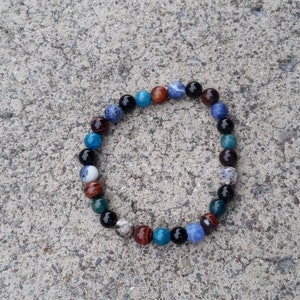 Puede incluir: Una pulsera de cuentas multicolores, incluyendo cuentas azules, negras, marrones y blancas. La pulsera circular descansa sobre una superficie gris texturizada. Las cuentas son redondas y pulidas.