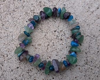 Può includere: Un braccialetto elastico composto da frammenti irregolari di pietre preziose levigate in varie tonalità di verde, blu e viola. Le pietre sono infilate in una forma circolare, mostrando un mix di consistenze traslucide e opache.