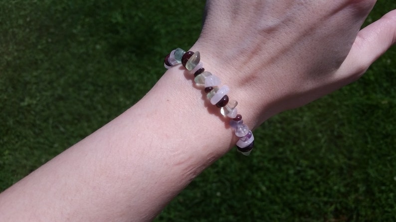 Op de afbeelding: Een armband gemaakt van groene, paarse en roze stenen. De armband zit om een pols.