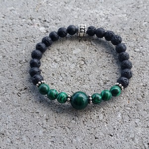 May include: A beaded bracelet featuring black lava rock beads and green malachite stones. The bracelet has a central malachite bead and silver-colored spacers. The bracelet is displayed on a gray surface.