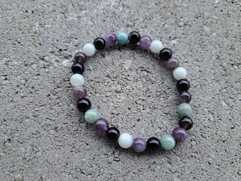 Pu&ograve; includere: Un braccialetto di perline composto da varie pietre preziose. Le perline rotonde sono di vari colori, tra cui nero, bianco, viola, verde e azzurro. Il braccialetto &egrave; appoggiato su una superficie grigia e strutturata.
