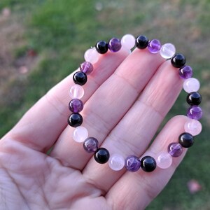 May include: A beaded bracelet featuring a mix of gemstones. The bracelet includes round beads in shades of black, purple, and pink. The bracelet is displayed against a blurred green background.