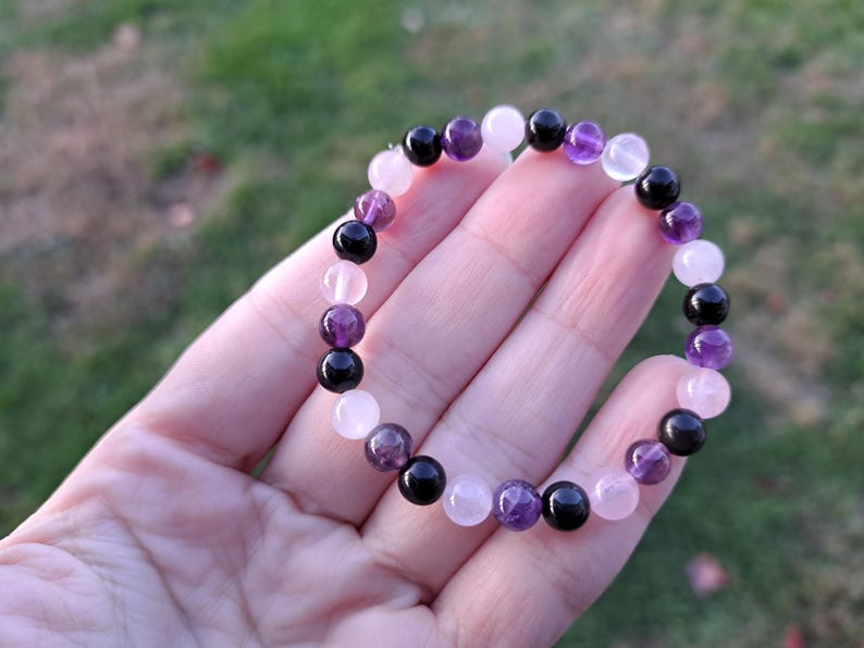 May include: A beaded bracelet featuring a mix of gemstones. The bracelet includes round beads in shades of black, purple, and pink. The bracelet is displayed against a blurred green background.