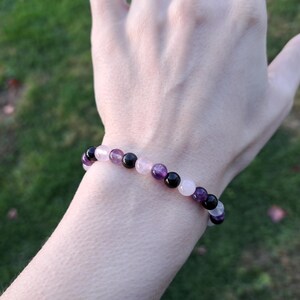 May include: A beaded bracelet featuring a mix of amethyst, rose quartz, and black onyx gemstones. The bracelet is strung on an elastic cord and is worn on a wrist. The background is a blurred view of green grass.