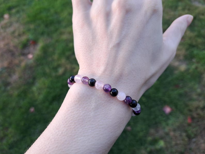 May include: A beaded bracelet featuring a mix of amethyst, rose quartz, and black onyx gemstones. The bracelet is strung on an elastic cord and is worn on a wrist. The background is a blurred view of green grass.