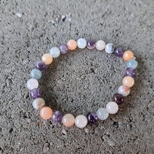 May include: A beaded bracelet composed of various colored gemstones, including purple, peach, white, and blue. The bracelet is circular and rests on a textured gray surface. The beads are round and appear to be polished.