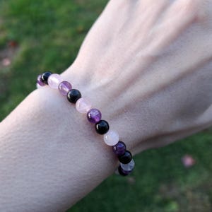 May include: A beaded bracelet featuring a mix of gemstones. The bracelet includes black, purple, pink, and white beads. The bracelet is worn on a wrist, with a blurred green background.