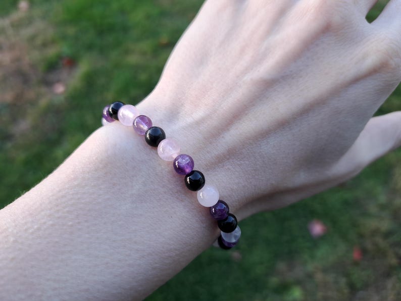 May include: A beaded bracelet featuring a mix of gemstones. The bracelet includes black, purple, pink, and white beads. The bracelet is worn on a wrist, with a blurred green background.