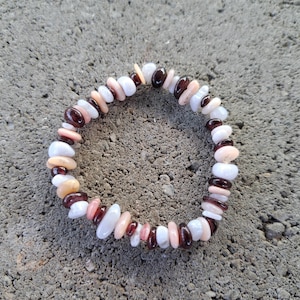 May include: A beaded bracelet composed of various natural stone chips. The bracelet features a mix of colors, including white, light pink, dark red, and tan. The stones are strung together to form a circular shape, suitable for wrist wear.