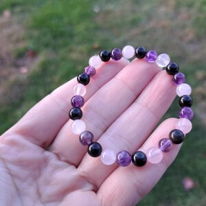May include: A beaded bracelet featuring a mix of amethyst, rose quartz, and black onyx gemstones. The bracelet is held in a hand against a blurred green background. The beads are approximately 0.3 inches in diameter.