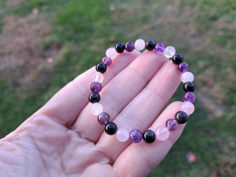 May include: A beaded bracelet featuring a mix of amethyst, rose quartz, and black onyx gemstones. The bracelet is held in a hand against a blurred green background. The beads are approximately 0.3 inches in diameter.