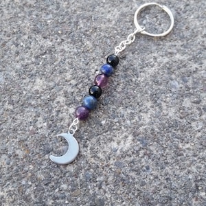 May include: A silver keychain with a crescent moon charm and a chain of colorful beads. The beads are purple, blue, and black. The keychain is on a gray, textured surface.
