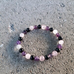 May include: A beaded bracelet featuring a mix of gemstones. The bracelet includes black, purple, pink, and white beads. The bracelet is circular and rests on a gray, textured surface.