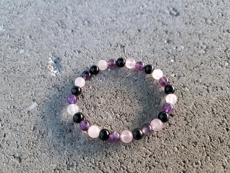 May include: A beaded bracelet featuring a mix of gemstones. The bracelet includes black, purple, pink, and white beads. The bracelet is circular and rests on a gray, textured surface.