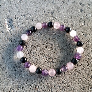 May include: A beaded bracelet featuring a mix of gemstones. The bracelet includes round beads in shades of black, purple, pink, and white. The bracelet is displayed on a gray, textured surface.