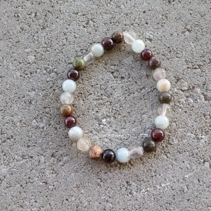 May include: A bracelet made of various colored stones, including brown, red, green, and white. The bracelet is made of round beads and is strung on a clear elastic cord.