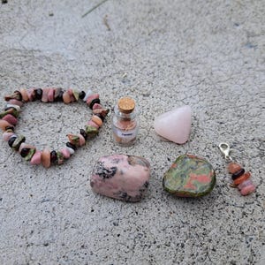 May include: A collection of gemstones and a bracelet. The bracelet is made of pink, green, and brown stones. There is a small glass bottle with a cork, a pink triangular stone, and a charm with multiple stones.