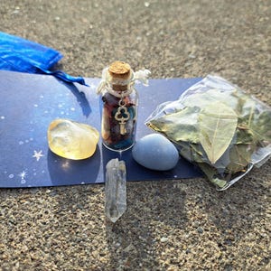May include: A small glass bottle filled with colorful crystals and a gold key charm. The bottle is sitting on a blue background with white stars. There is a clear quartz crystal, a yellow stone, and a gray stone next to the bottle. A bag of dried bay leaves is in the background.