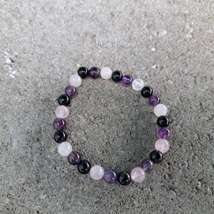 May include: A beaded bracelet composed of various gemstones, including black, purple, and pink beads. The bracelet is made of round beads, and the colors are arranged in a repeating pattern. The bracelet is resting on a gray surface.