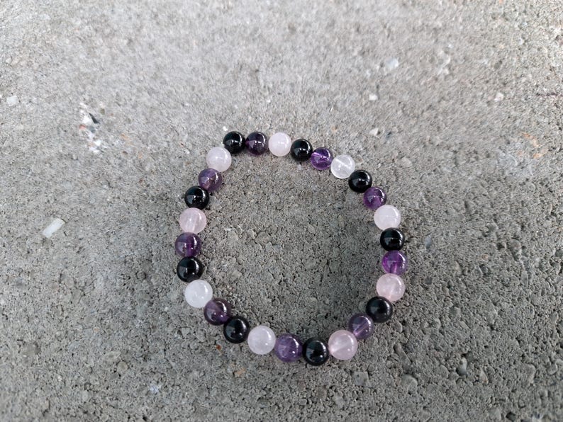 May include: A beaded bracelet composed of various gemstones, including black, purple, and pink beads. The bracelet is made of round beads, and the colors are arranged in a repeating pattern. The bracelet is resting on a gray surface.