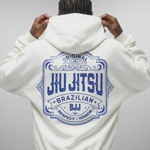 Puede incluir: Sudadera con capucha color crema con un gráfico azul en la espalda. El gráfico dice "ORIGINAL JIU JITSU BRAZILIAN BJJ RESPECT HONOR" en un marco decorativo. La sudadera tiene capucha y mangas largas.