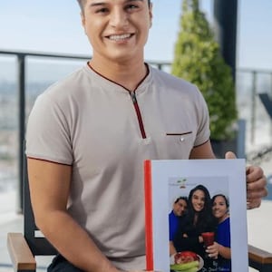 以下が含まれることがあります： 赤い背表紙と表紙に写真が付いた本を持っている人。写真には3人の人物と料理の盛り合わせが写っています。その人は、バーガンディのジッパーが付いた明るい灰色のシャツを着ています。