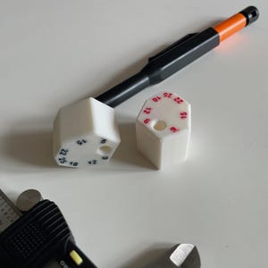以下が含まれることがあります： 上面に赤色で6、9、10、12、15、22、25の数字が印刷された、2つの白い六角形のプラスチック製コンポーネント。黒とオレンジのシャープペンシルと、黒いデジタルノギスが、2つの白い物体の横に配置されています。