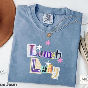 May include: A blue jean coloured t-shirt featuring a graphic print with the text Lunch Lady in a collage style font. The design includes small pink star shapes and white star shapes with black dots. The shirt has a crew neckline and a Comfort Colors label.