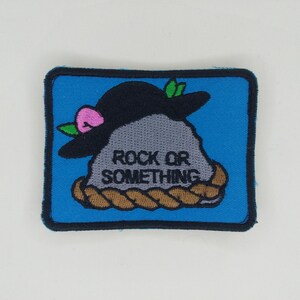 May include: A rectangular patch with a blue background and a black border. The patch features a gray rock wearing a black hat with a pink flower and green leaves. The text "ROCK OR SOMETHING" is below the rock, and a brown rope design is at the bottom.