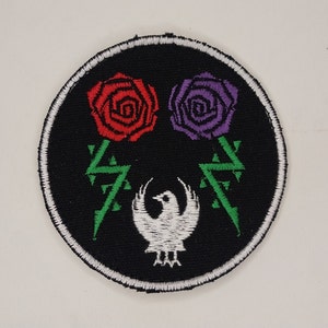 May include: Embroidered patch with a black background, featuring a red rose and a purple rose above a white phoenix. Green lightning bolt-like designs flank the roses. The patch has a white stitched border.