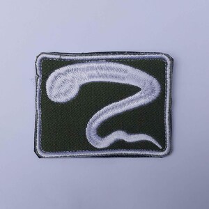 May include: Embroidered patch featuring a white, stylized snake design on a dark green rectangular background. The patch has a white border and is likely for clothing or accessories.