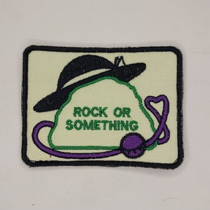 May include: A green and purple embroidered patch with a black hat on top of a rock. The text "Rock or Something" is embroidered in green on the patch.