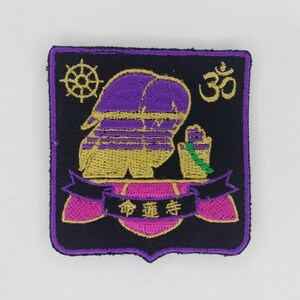 May include: Purple and gold embroidered patch with a figure in a prayer pose, a green mala, and the word "Om" in gold. The patch has a pink banner with the text "生命" in gold.