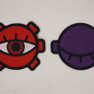 May include: Two embroidered patches, one red with a black eye and the other purple with a black eye. Both patches have a black outline.