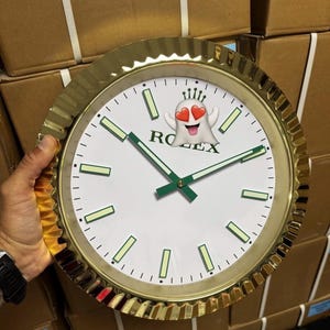 Puede incluir: Un reloj de pared redondo con un bisel estriado de color dorado y una esfera blanca. La esfera presenta marcadores de hora y manecillas luminiscentes verdes, con el texto ROLEX impreso en negro. Un emoji de fantasma con ojos de corazón lo cubre.