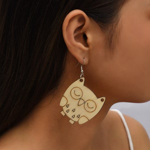 Könnte beinhalten: Ein Paar hölzerne Eulenohrringe mit einer natürlichen Holzoptik. Die Ohrringe sind in Form von Eulen mit geschlossenen Augen und einem kleinen Schnabel geformt. Die Ohrringe sind an silbernen Haken befestigt.