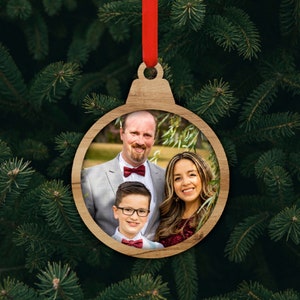 May include: Wooden Christmas ornament in the shape of a ball, with a family photo inside. The ornament is suspended by a red ribbon against a backdrop of green pine needles. The family is smiling and wearing formal attire.