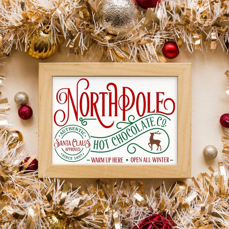 May include: A framed sign with the text "NORTH POLE HOT CHOCOLATE CO." in red and green lettering. The sign also includes the text "AUTHENTIC. SANTA CLAUS APPROVED. SINCE 1897." and a small image of a reindeer.