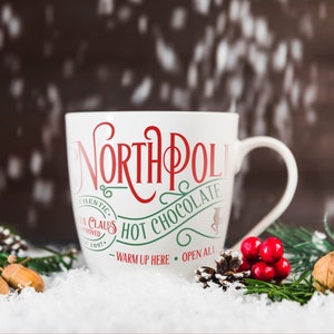 May include: White ceramic mug with red and green text that reads "North Pole Hot Chocolate - Warm Up Here - Open All". The mug is sitting on a snowy surface with pine branches and acorns.