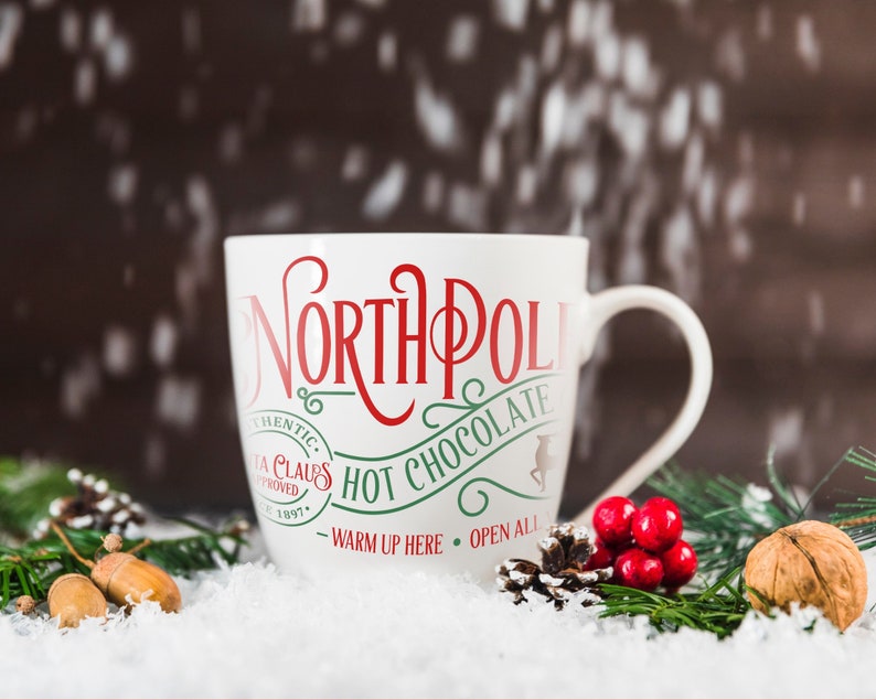 May include: White ceramic mug with red and green text that reads "North Pole Hot Chocolate - Warm Up Here - Open All". The mug is sitting on a snowy surface with pine branches and acorns.