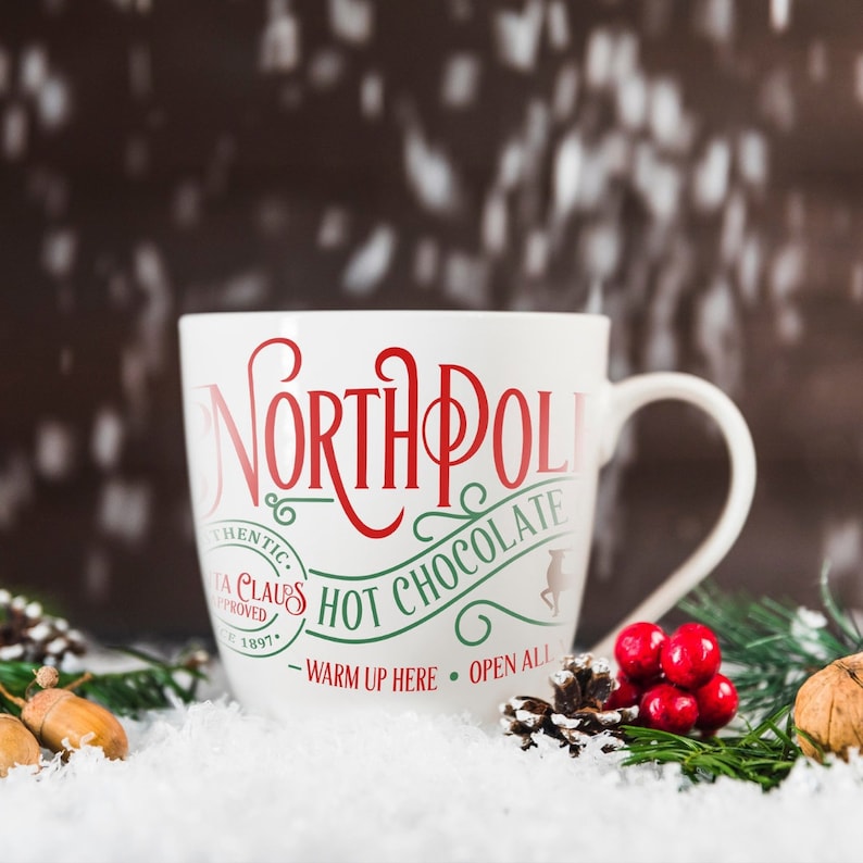 May include: White ceramic mug with red and green text that reads "North Pole Hot Chocolate - Warm Up Here - Open All". The mug is sitting on a snowy surface with pine branches and red berries.