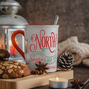 May include: White ceramic mug with a red handle and green and red text that reads "North Pole Santa Claus Hot Chocolate Warm Up Here". The mug is sitting on a wooden surface with a pine cone, star anise, and cookies.