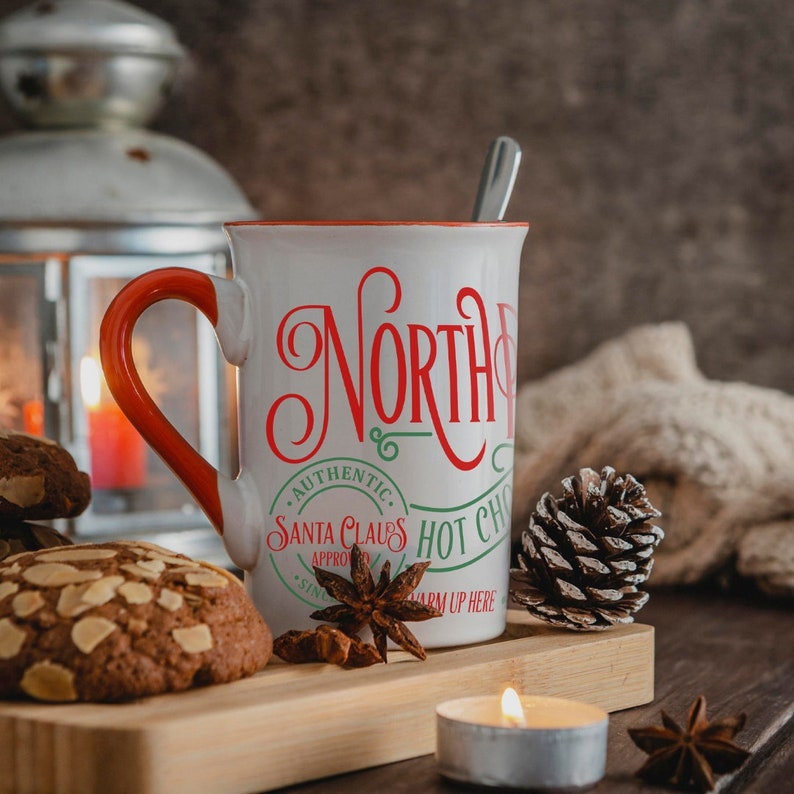 May include: White ceramic mug with a red handle and green and red text that reads "North Pole Santa Claus Hot Chocolate Warm Up Here". The mug is sitting on a wooden surface with a pine cone, star anise, and cookies.